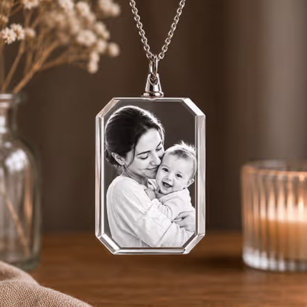 Rechteckige 2D-Kristall-Halskette mit eingraviertem Porträt einer Mutter mit ihrem Baby, an einer Silberkette neben einer leuchtenden Kerze.