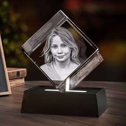 Diamantförmiger 3D-Fotokristall mit eingraviertem Porträt eines lächelnden jungen Mädchens, präsentiert auf einem schwarzen LED-Lichtsockel neben dem Originalfoto.