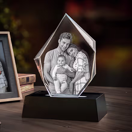 Isbergförmiger 3D-Fotokristall mit eingraviertem Porträt von Eltern und ihrem Baby, präsentiert auf einem schwarzen LED-Lichtsockel neben dem Original-Familienfoto.