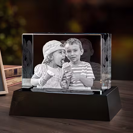 Breiter rechteckiger 3D-Fotokristall mit eingraviertem Porträt von zwei Kindern, die sich ein Eis teilen, präsentiert auf einem schwarzen LED-Lichtsockel neben dem Originalfoto.